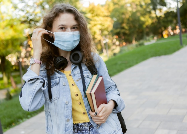 Curly hair woman student in facial medical mask holding a stack of books outdoors Education during a pandemic