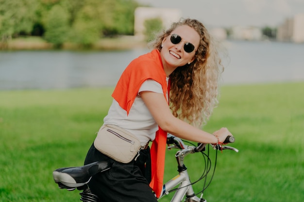 Curly haired blonde woman cycles outdoor stands with bicycle carries bag wears sunglasses poses against green lawn wears white t shirt black trousers red sweater on shoulders Outdoor lifestyle