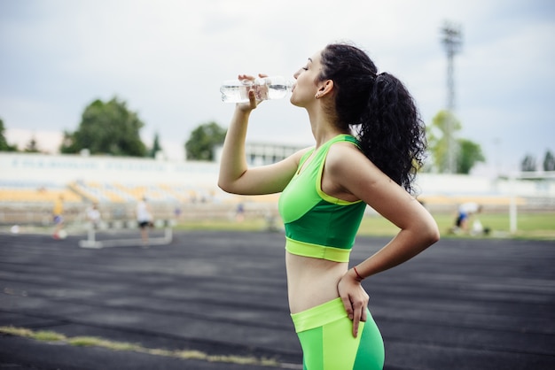 Curly-haired brunette playing sports at the stadium. Girl drinks water from a bottle