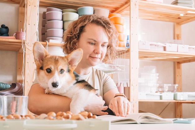 Curly haired glad cheerful businesswoman owner with dog corgi sitting relaxing and reading book at workplace indoors Small business silicone dishes for babies Home comfortable office store