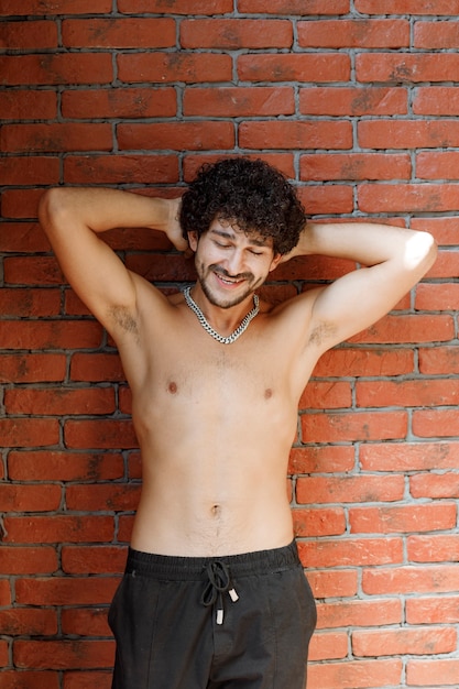 Curly haired half naked man posing to camera against wall.