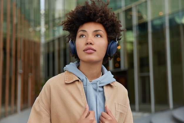 Curly haired teenage girl listens music via headphones focused somewhere with pensive expression dressed in hoodie and jacket strolls outdoors poses against blurred background Lifestyle concept