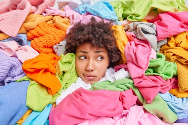 Photo curly haired woman collects clothes items in good condition to consignment shop or thrift store surrounded by huge piles of multicolored garments focused away has tired expression. recycling textiles