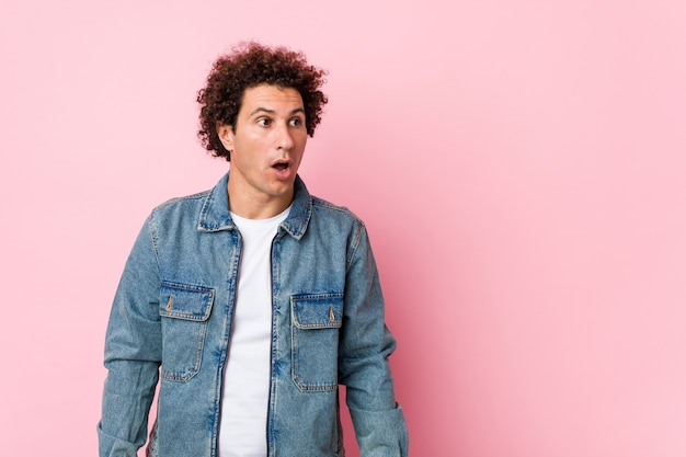 Curly mature man wearing a denim jacket against pink  being shocked because of something she has seen.