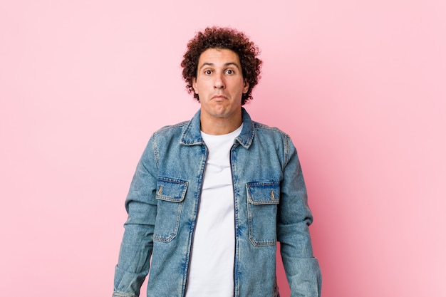 Curly mature man wearing a denim jacket against pink wall shrugs shoulders and open eyes confused.