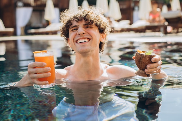 Curly positive guy holds cocktails and enjoys spending time in the pool