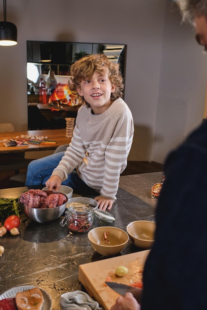 Curly smiling boy in sweater sitting on wide kitchen counter full of kitchenware against father