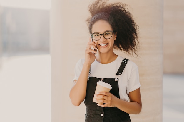 Curly teenage girl wears transparent glasses, talks on mobile phone, has coffee break, dressed in white t shirt