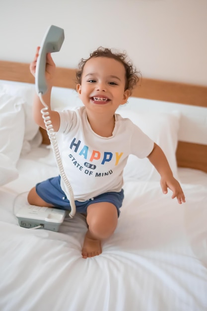 Curly toddler boy playing with a wired telephone on the bed