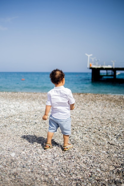 Curly toddler boy plays on the pebble beach near blue sea