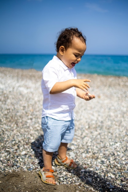 Curly toddler boy plays on the pebble beach near blue sea