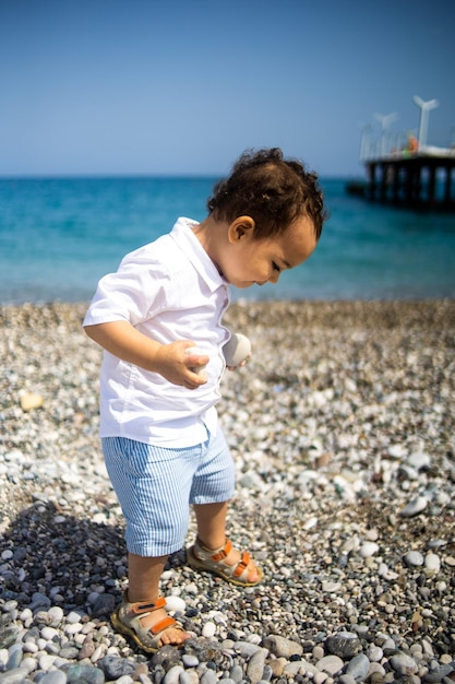 Curly toddler boy plays on the pebble beach near blue sea