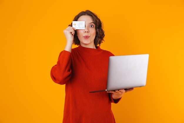 curly woman wearing sweater using silver laptop and credit card while standing isolated on yellow