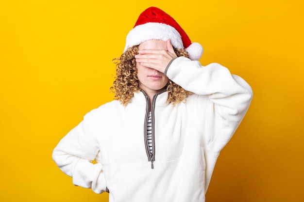 Curly young woman in a santa hat with palms closed her eyes on a yellow background.