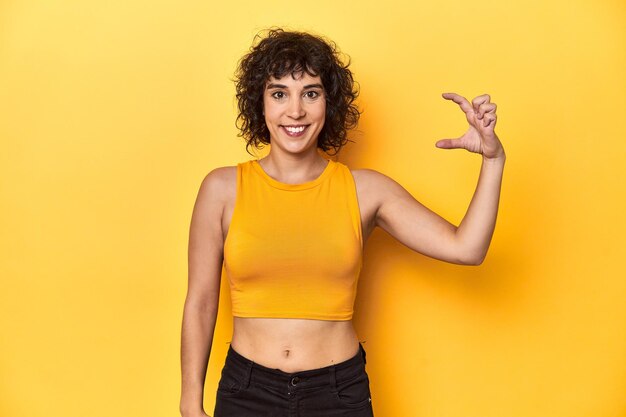 Curlyhaired Caucasian woman in yellow top holding something little with forefingers smiling