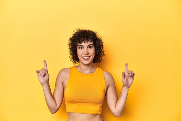 Photo curlyhaired caucasian woman in yellow top indicates with both fore fingers up showing a blank space