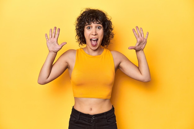 Photo curlyhaired caucasian woman in yellow top receiving a pleasant surprise excited and raising hands