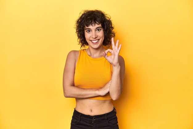 Photo curlyhaired caucasian woman in yellow top winks an eye and holds an okay gesture with hand