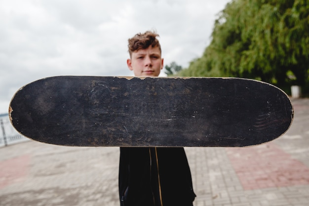 A curlyhaired european teen holds a black old shabby skate in front of him only the skate is in focu...