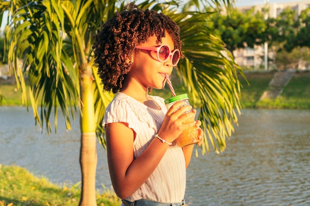 Curlyhaired girl drinks juice from bottle through straw while walking outdoors