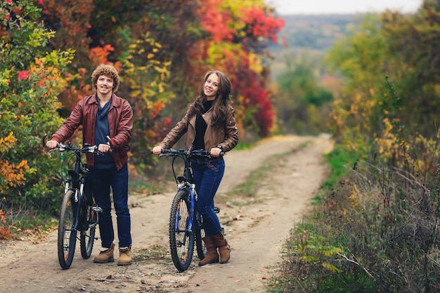 Curlyhaired mustachioed guy and an orehaired girl in autumn stand with bicycles on road and show cheerful emotions