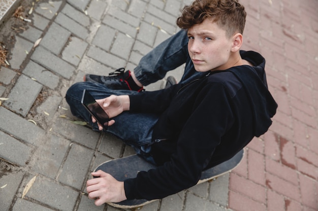 A curlyhaired teen of european appearance in black hoodie sitting on an alley on a skateboard and ta...