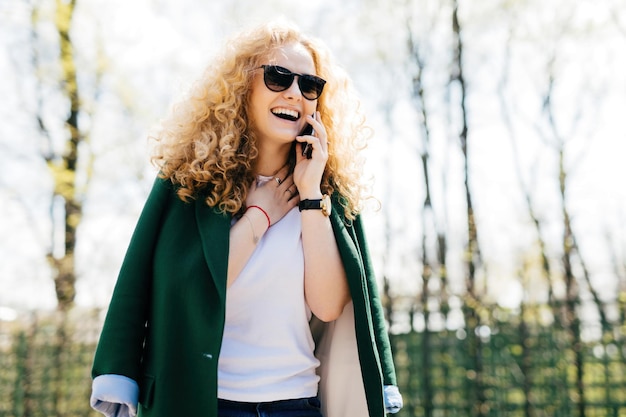 Curlyhaired woman in stylish clothes and sunglasses talking on smartphone smiling and laughing with excitement Happy expression neck touch