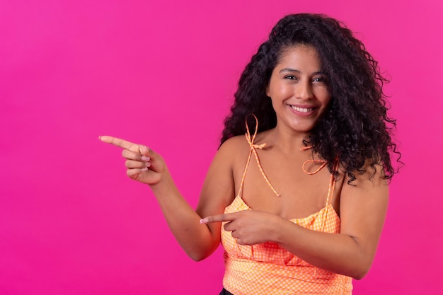 Curlyhaired woman in summer clothes on a pink background pointing left studio shot