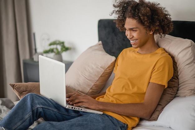 Curlyhaired young guy sitting on a bed with a laptop