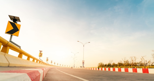 Curve concrete road with curve traffic sign beside the sea at sunset time. Solar panel energy on yellow curve traffic sign. Road trip on summer vacation. Blur car driving. Summer travel by car.