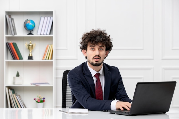 Customer service curly brunette young man in office suit and red tie with laptop typing