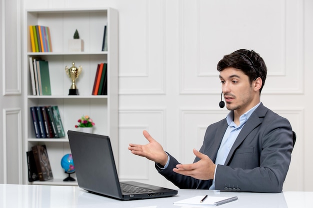 Customer service cute guy in grey suit with computer and headset having a videocall