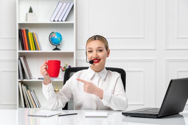 Customer service pretty blonde girl in white shirt with laptop and headset pointing at red cup