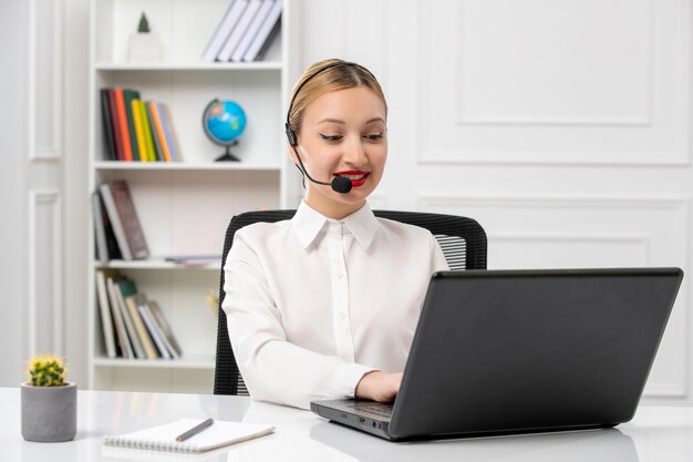 Customer service pretty blonde girl in white shirt with laptop and headset typing and smiling