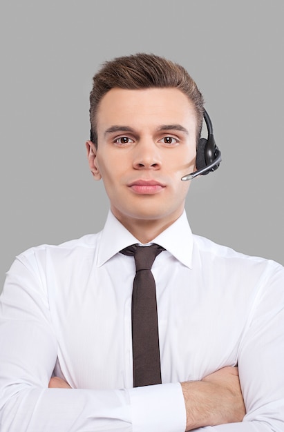 Customer service representative. Cheerful young man in formalwear adjusting his headset and smiling at camera while standing against grey background