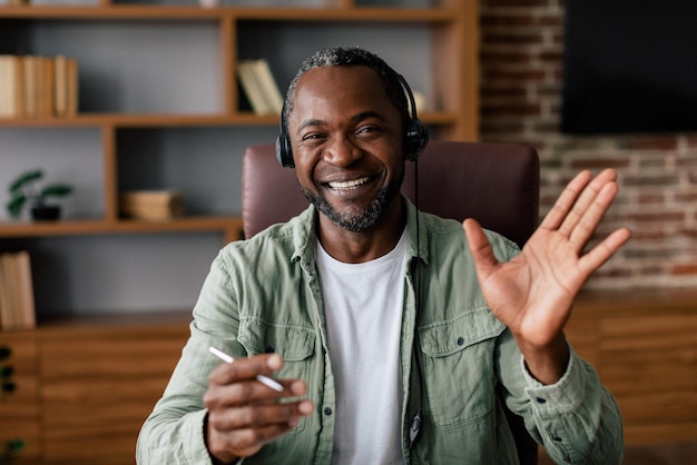 Customer support and work at home Smiling adult african american guy manager in casual and headphones waves hand