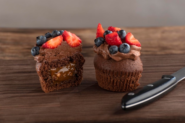 Cut chocolate cupcake is decorated with chocolate butter cream and berries Dessert and knife on rough wooden brown board