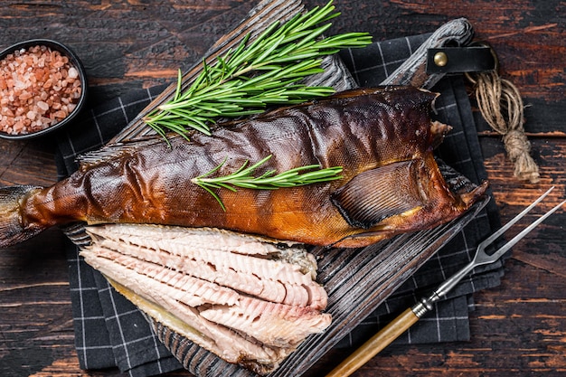 Cut Hot smoked fish  on a wooden board with rosemary. Dark wooden background. Top view.