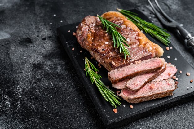 Cut roasted new york strip beef meat steak or striploin on a marble board. Black background. Top view. Copy space.