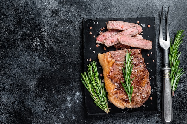 Cut roasted new york strip beef meat steak or striploin on a marble board. Black background. Top view. Copy space.