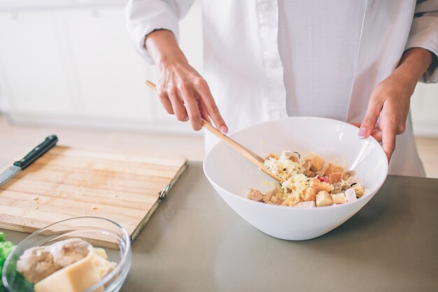 Cut view of girl's hands mixing salad in white bowl. She is using spoon for that. Girl wears white shirt. She cooks.