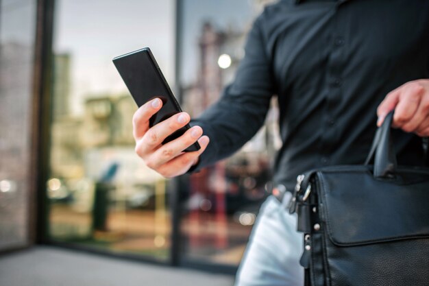 Cut view of man stand at building outside. He hold phone and bag.