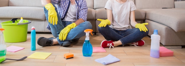 Cut view of man and woman sitting on the floor and resting. She is meditating. Man and woman has their legs crossed. There are cleaning equipment all over the floor.