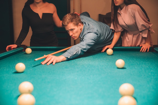 Cut view of young man looking concentrated. He aiming into billiard ball. Two slim models stand behind him.