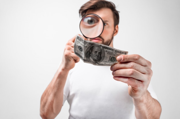 Cut view of a young man that holds a hundred dollar bill and looking on it through the loupe. He wants to be sure that this bill is not a fake bill. Isolated on white wall