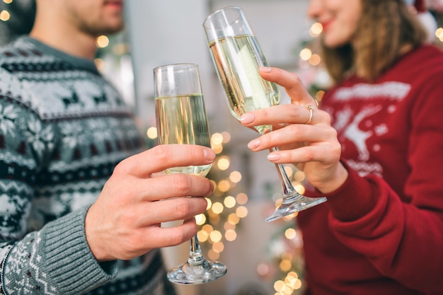 Cut view of young man and woman standing in front of each other. They wear Christmas sweaters. Couple holds glasses of champaigne. She smiles.