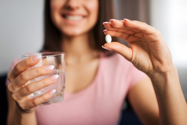 Cut view of young positive woman hold white pill and glass of water. She smiles to camera.