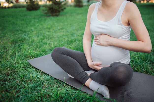 Cut view of young pregnant woman sit on yoga mate in park. She hold hands around belly. Model sit in lotus pose.