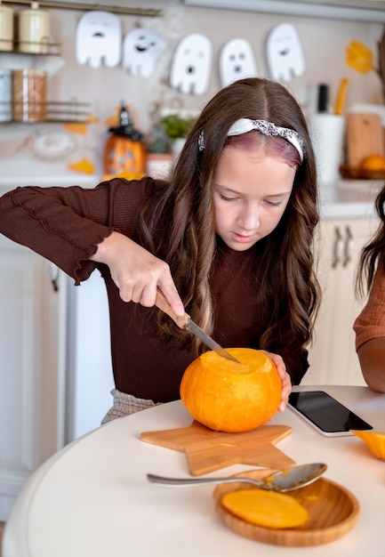 Cute 10 years old girl holding knife ang carving spooky face on pumpkin in light white kitchen at home Halloween preparation