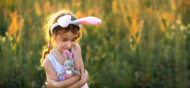 Cute 5-year-old girl with rabbit ears gently hugs a toy rabbit in nature in a blooming field in summer with golden sunlight. Easter, Easter bunny, childhood, happy child, springtime.